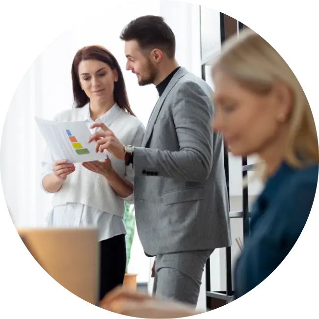 A man in a gray suit and a woman in a white blouse review a document with colorful charts, possibly discussing ServiceNow FSM, while a blonde woman in the foreground works on her computer.